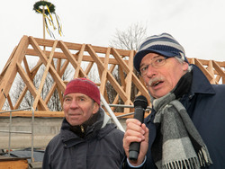 Auf einer Balkenkonstruktion ragt ein Richtkranz, vor dem Gebäude stehen zwei Männer. 